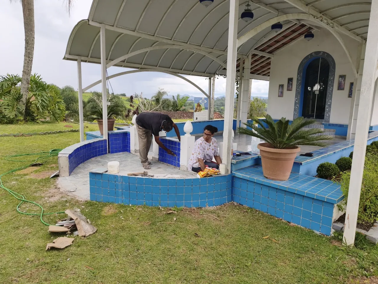 Pessoas trabalhando na manutenção de uma varanda com colunas brancas e revestimento azul, cercada por jardins.
