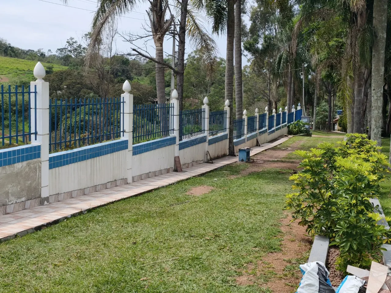 Caminho junto a um jardim, palmeiras e uma grade azul com pilares brancos.